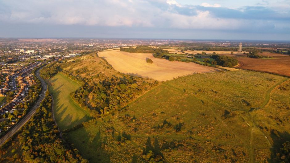 Aerial view of a rural landscape featuring a narrow, winding road cutting through dense green woodland, with open fields and farmland in the distance under a partly cloudy sky. The scene includes patc
