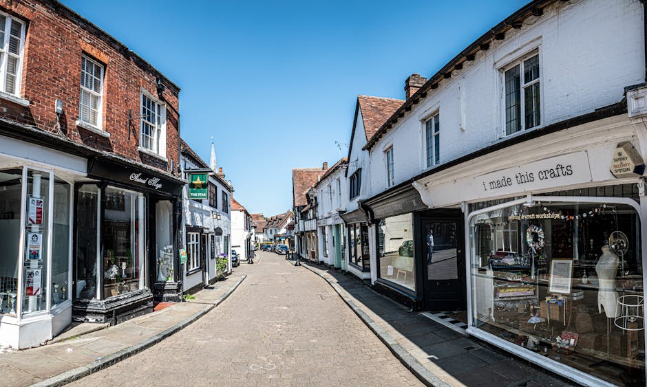 A narrow street in Guildford with small retail shops on both sides, showcasing a mix of brick and white-painted facades. The shopfronts include display windows filled with various items, such as decorative objects and jewelry, with one shop having signage indicating handmade crafts. The street is paved with cobblestones and appears quiet, with no pedestrians visible. Bright sunlight illuminates the scene, casting minimal shadows. In the background, residential buildings with tiled roofs are seen, and a green business sign hangs from one of the shops. The setting suggests a typical historic town centre suitable for house removals or furniture transport, and is relevant for visual content related to packing, loading, and home relocation services by Movers Surrey for their Guildford removals project for Cathedral and High Street moves, Surrey.