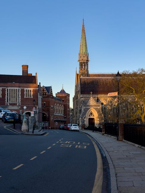 A street scene in Guildford during daytime with a clear blue sky, showing a narrow road with parked cars on the left side and a paved sidewalk on the right. In the background, a historic brick church with a tall, pointed, greenish spire is visible, along with other traditional red-brick buildings lining the street. The road features a dashed white line and a 'BUS STOP' marking on the pavement. A black streetlamp is positioned on the sidewalk near the church, and trees with partial foliage are visible on the right side of the image. The scene depicts an area suitable for house removals and furniture transport, with the architecture indicating central Guildford, supporting professional relocation and packing activities by Movers Surrey.